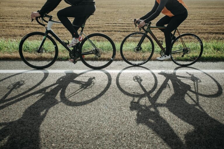 To cyklister cykler på en vej på deres racercykler med landevejsdæk ved dagslys. Deres skygger kastes ind over vejen.